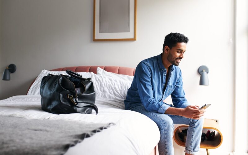Man Staying In Boutique Hotel Sits On Edge Of Bed Checking Emails And Social Media In The Morning Male guest in boutique hotel sitting on edge of bed checking emails and social media on mobile phone in the morning